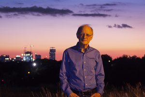 A man stands in a field at dusk with the city skyline behind him.